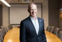 Caucasian man standing in front of a large conference table wearing a suit jacket and a shirt with no tie.