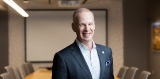 Caucasian man standing in front of a large conference table wearing a suit jacket and a shirt with no tie.